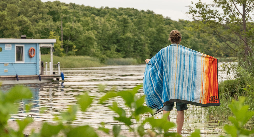 Person mit Klimahandtuch über die schultern gehangen steht mit den Füßen in einem grün umwachsenen See auf dem ein kleines Hausboot schwimmt