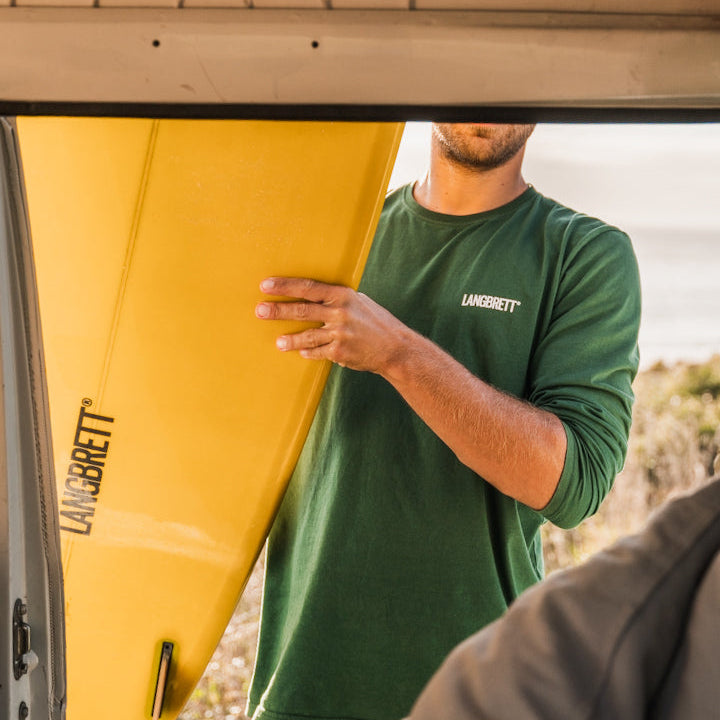 Person vor einem Auto in einem waldgrünen Langarmshirt mit einem sonnen gelbem Shortboard 